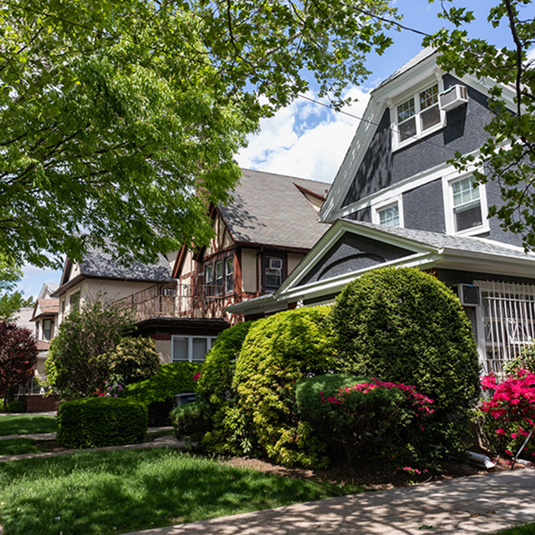 A black and white two-story house with a red brick chimney, situated on a tree-lined street, with a neatly manicured lawn and a clear blue sky overhead.