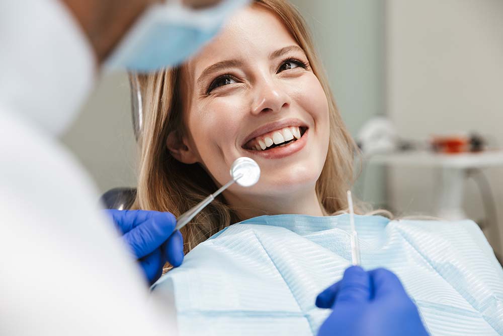 A woman is seated in a dental chair, smiling, while receiving dental treatment from a professional who is preparing her teeth with a dental drill.